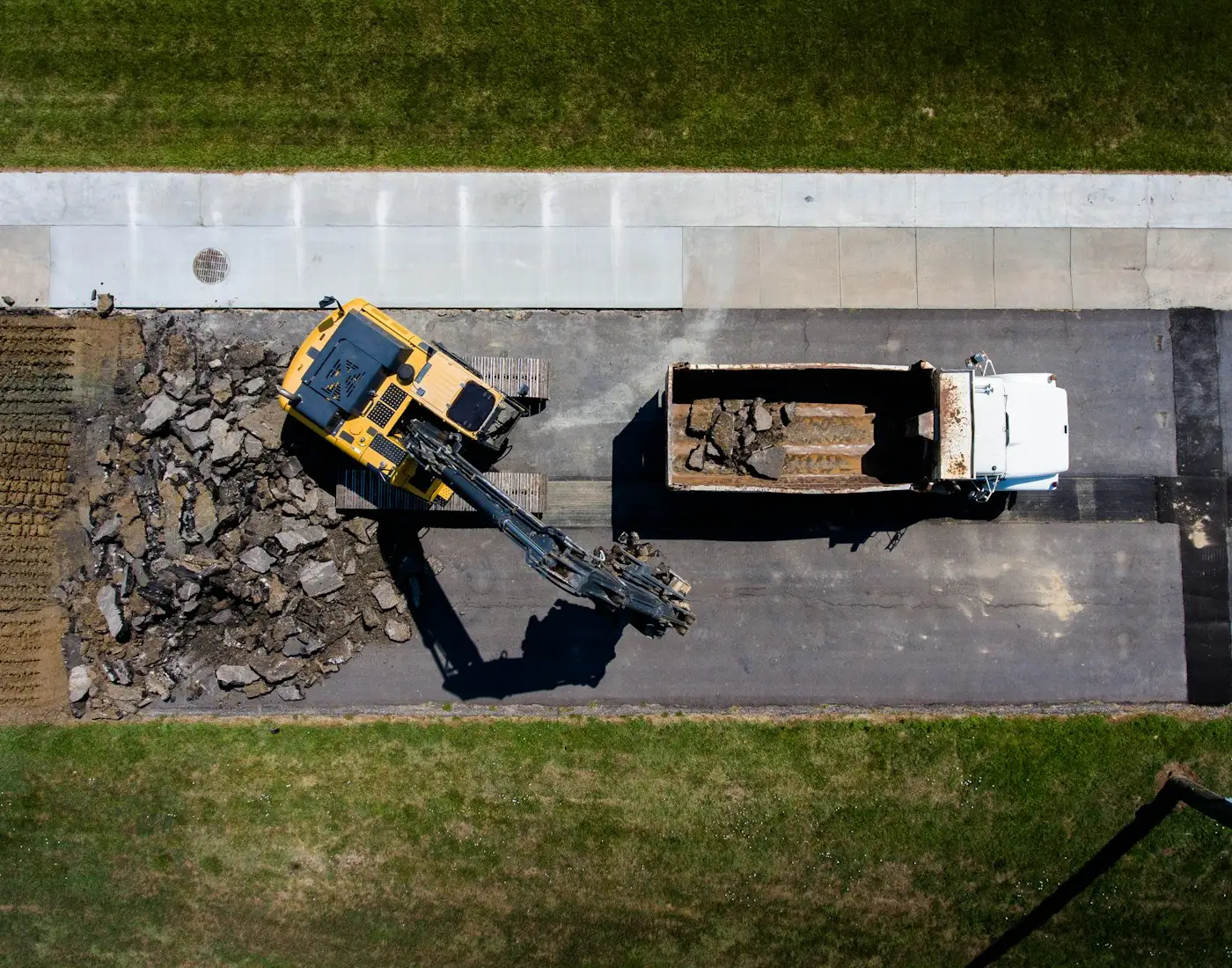 An excavator breaking out an old road surface and loading a tipper, seen from above
